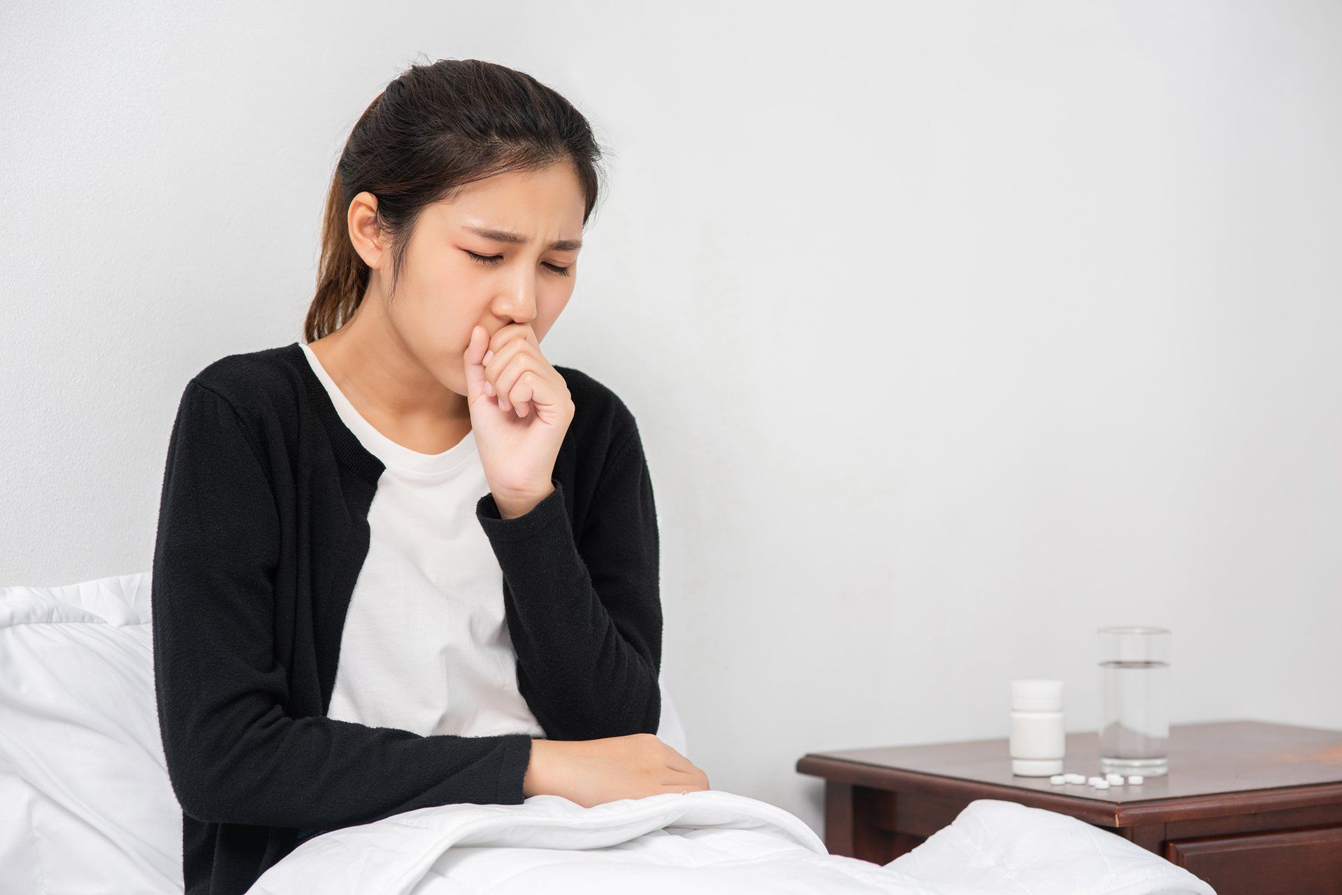 Woman coughing, covering her mouth with her hand, sitting on a bed next to a table with medicine and water.