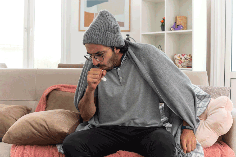 Man coughing, wrapped in a blanket, wearing a hat, sitting on a couch.