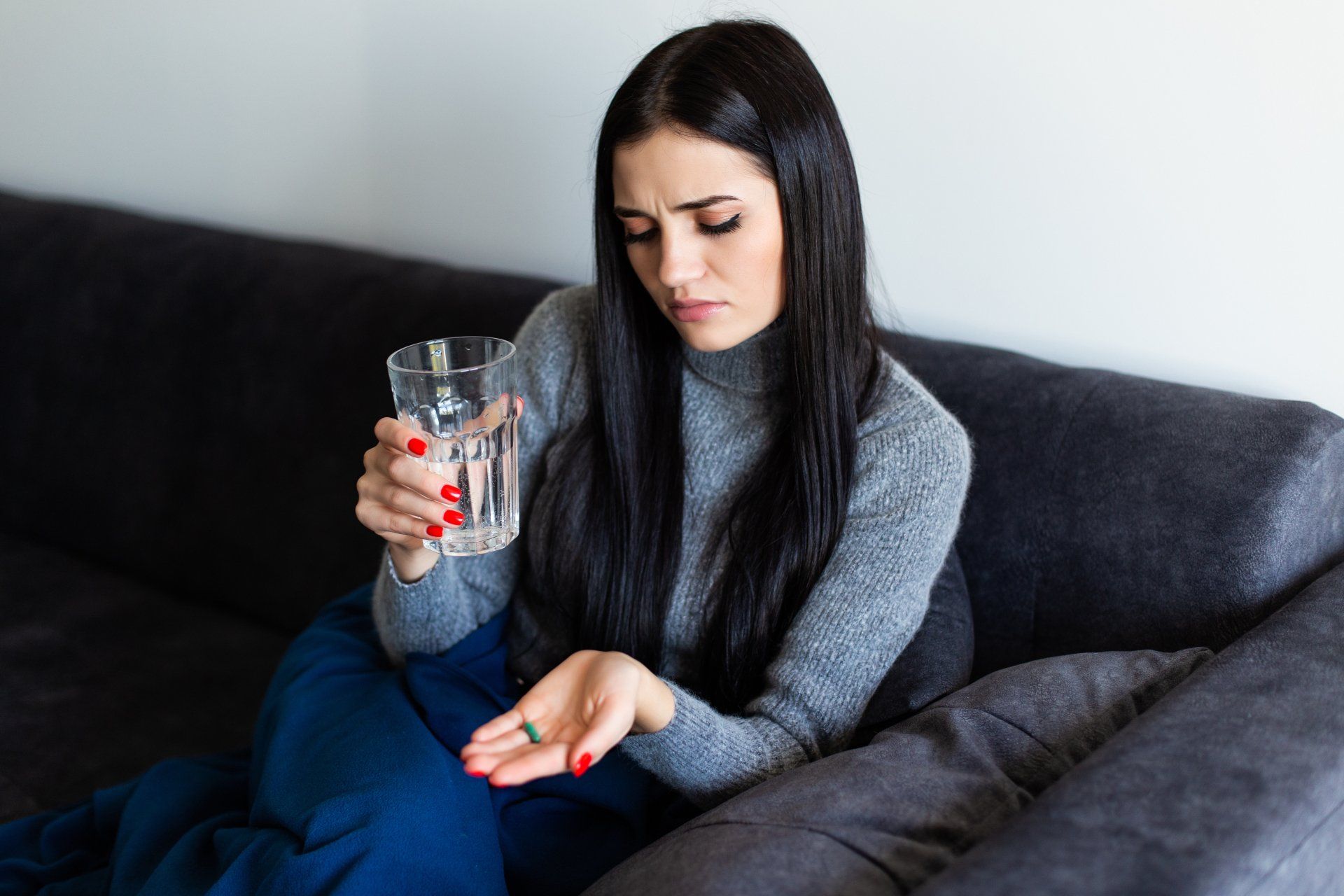 Woman on a sofa holding a pill and glass of water, looking distressed.