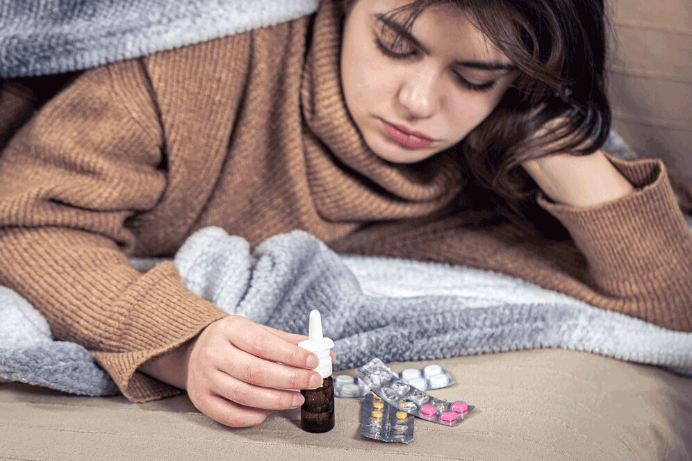Woman in bed holding nasal spray, with pills, looking unwell.