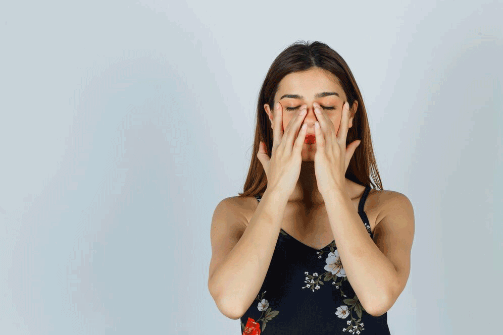 Woman with long brown hair, eyes closed, hands covering face. Wearing floral top, neutral background.