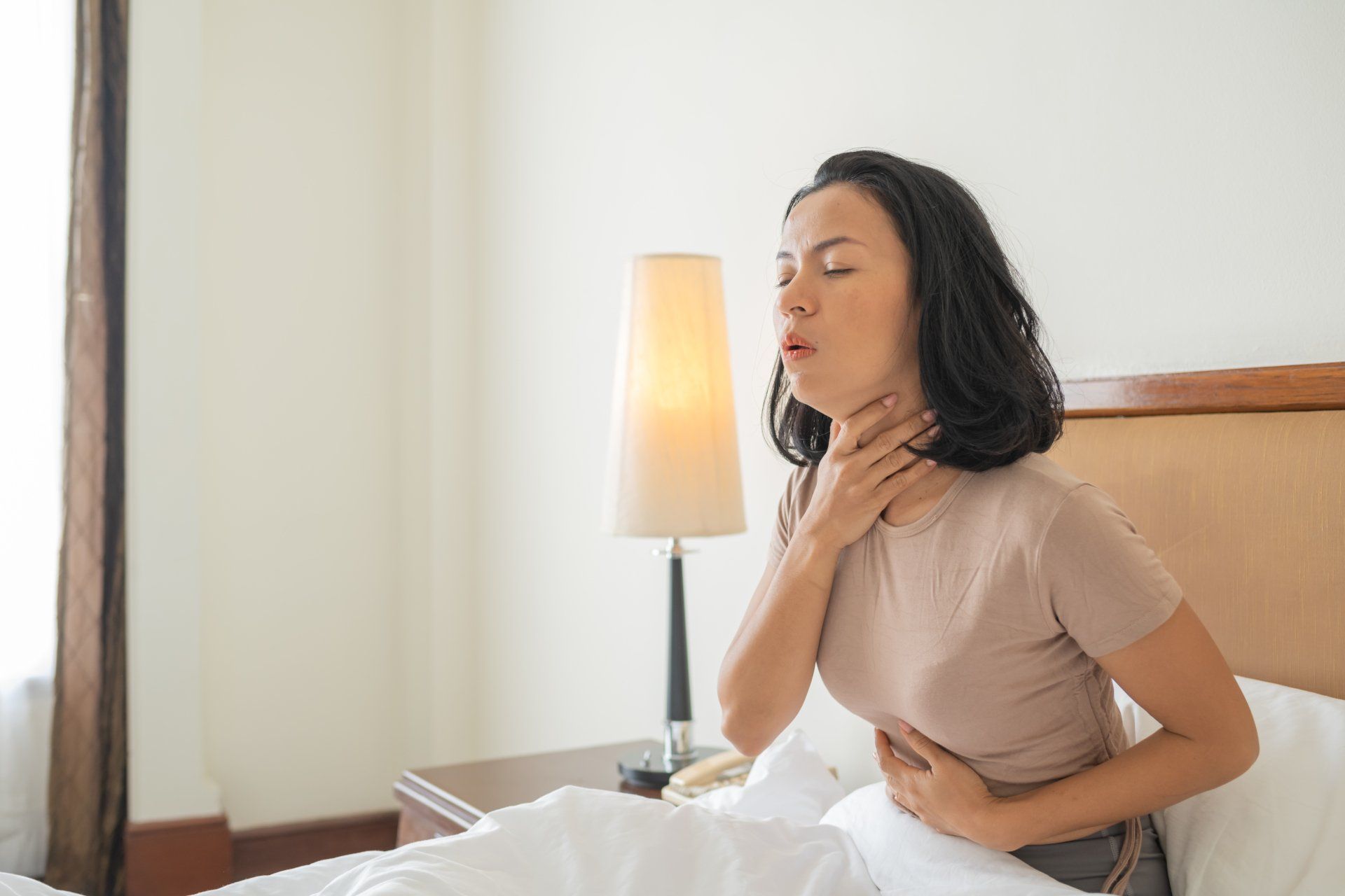 Woman sitting in bed, holding her neck and stomach, coughing, light room.