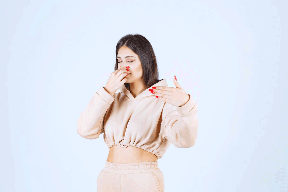 Woman in beige sweatsuit pinching her nose, gesturing away from herself; pale blue background.