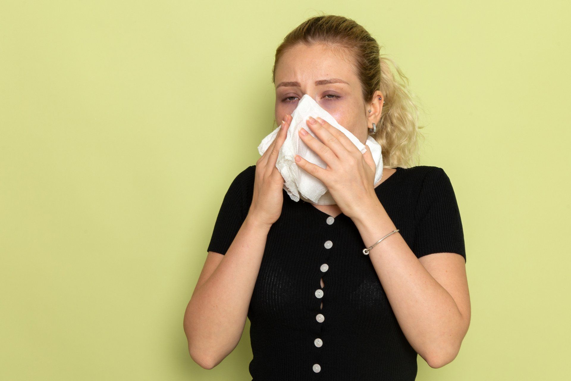 Woman with curly blonde hair blowing her nose into a white tissue, set against a light green background.