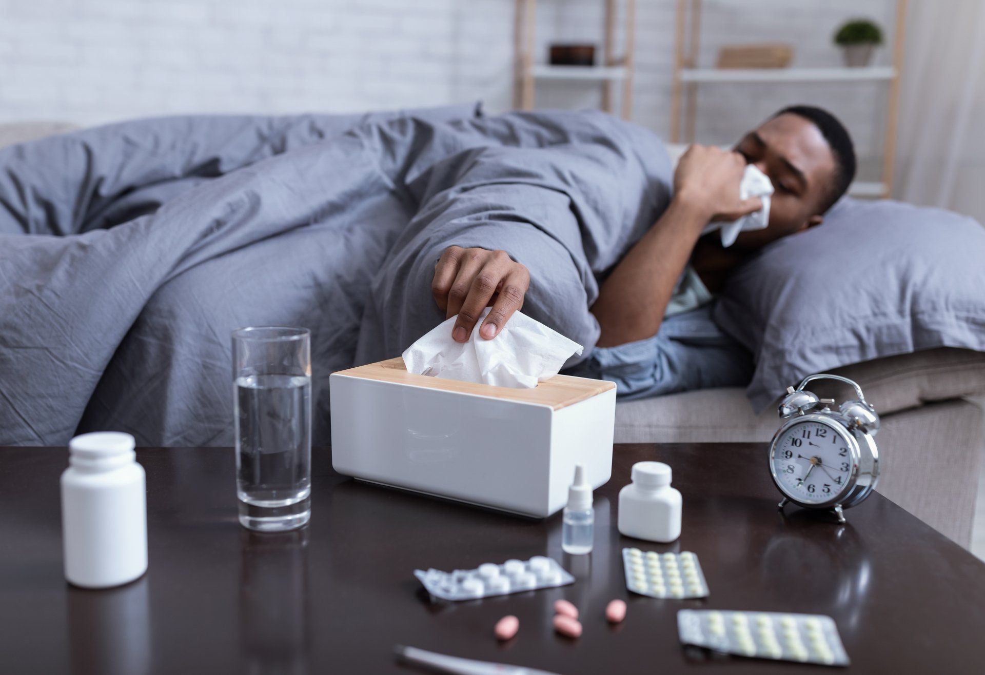 Person in bed blowing nose, reaching for tissues. Sickroom scene with pills, water, and clock.