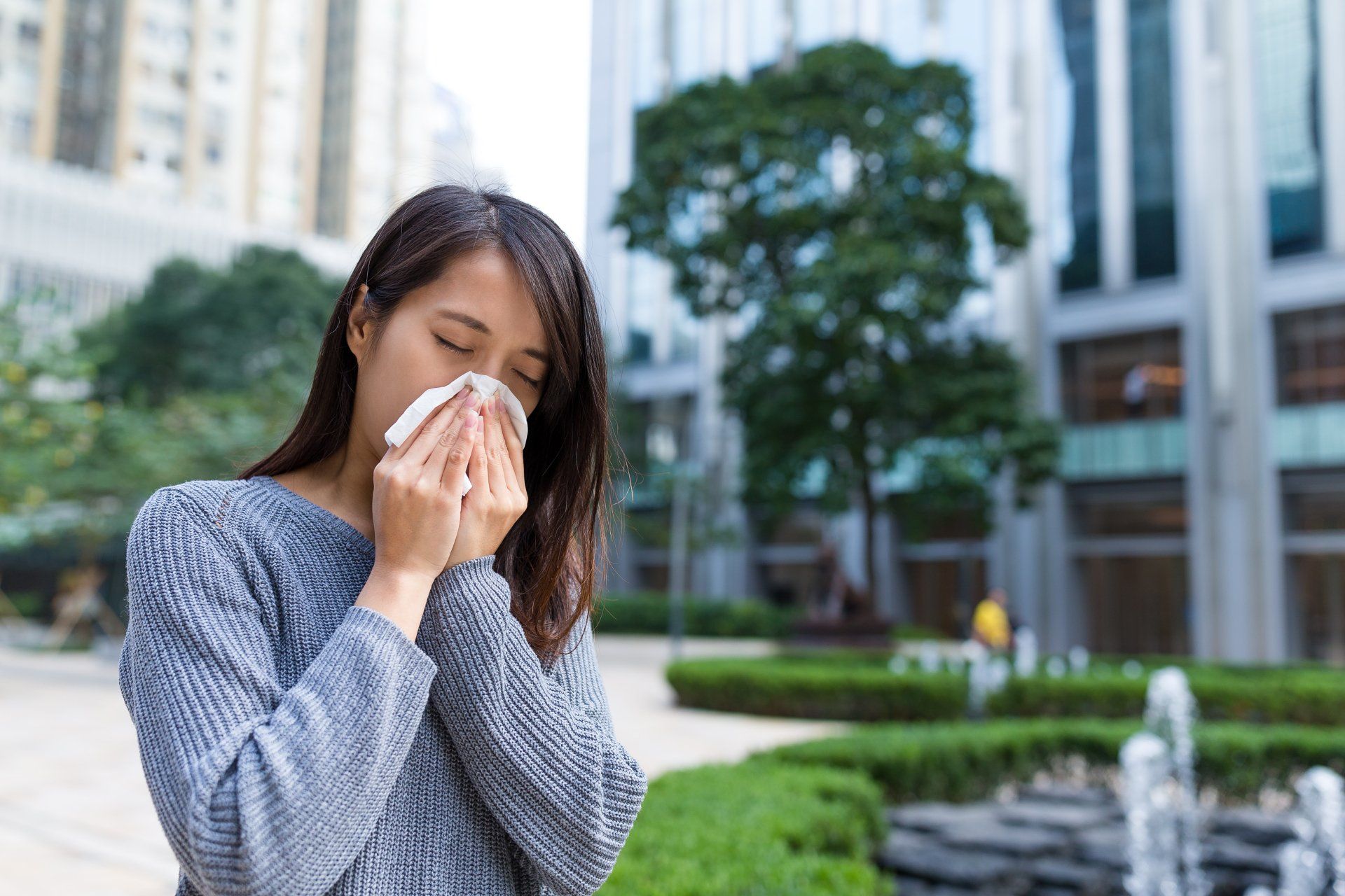 Woman sneezing into a tissue outdoors in a city, near greenery and buildings.
