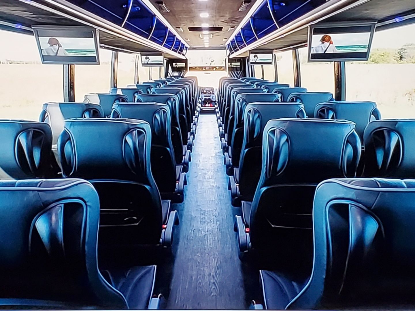 The inside of a bus with blue seats and televisions on the ceiling
