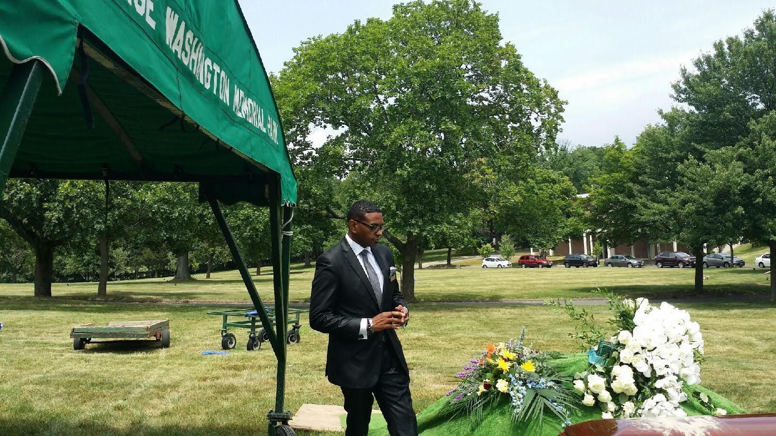 Man in black suit standing beside a gravesite with flowers under a green tent in a cemetery park