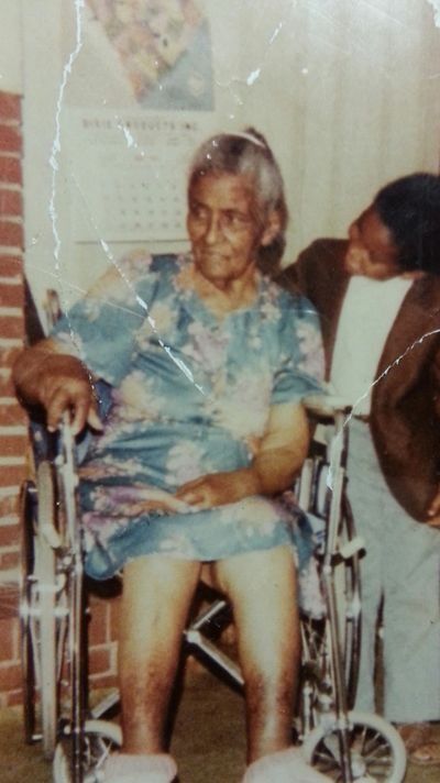 Elderly person seated in a wheelchair, wearing a blue shirt, with another person leaning in beside them.