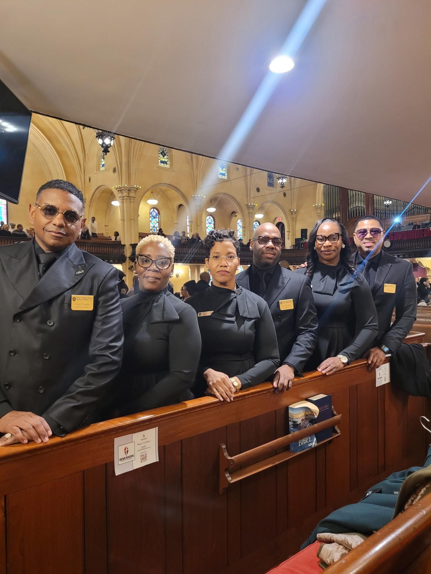 Uniformed group posing behind a wooden railing in a formal indoor setting, some holding railings and facing the camera