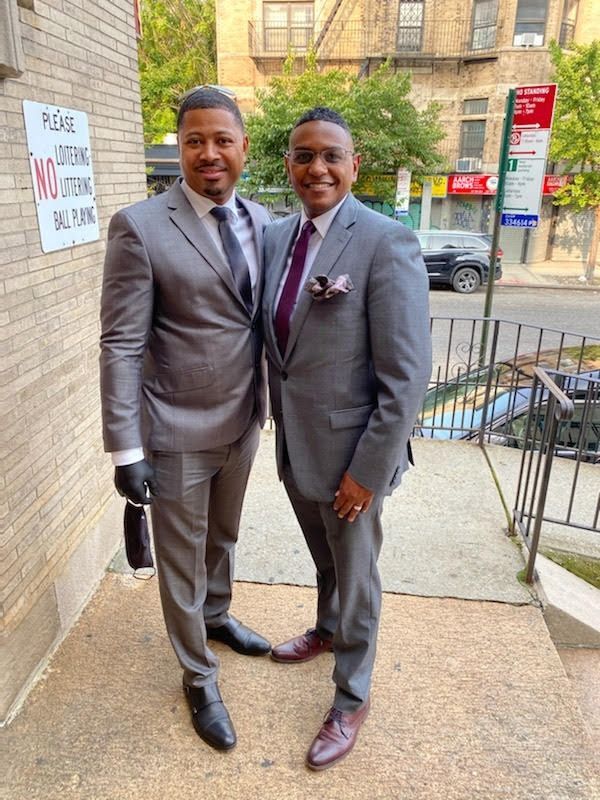 Two men in gray suits stand smiling on a city sidewalk outside a brick building.