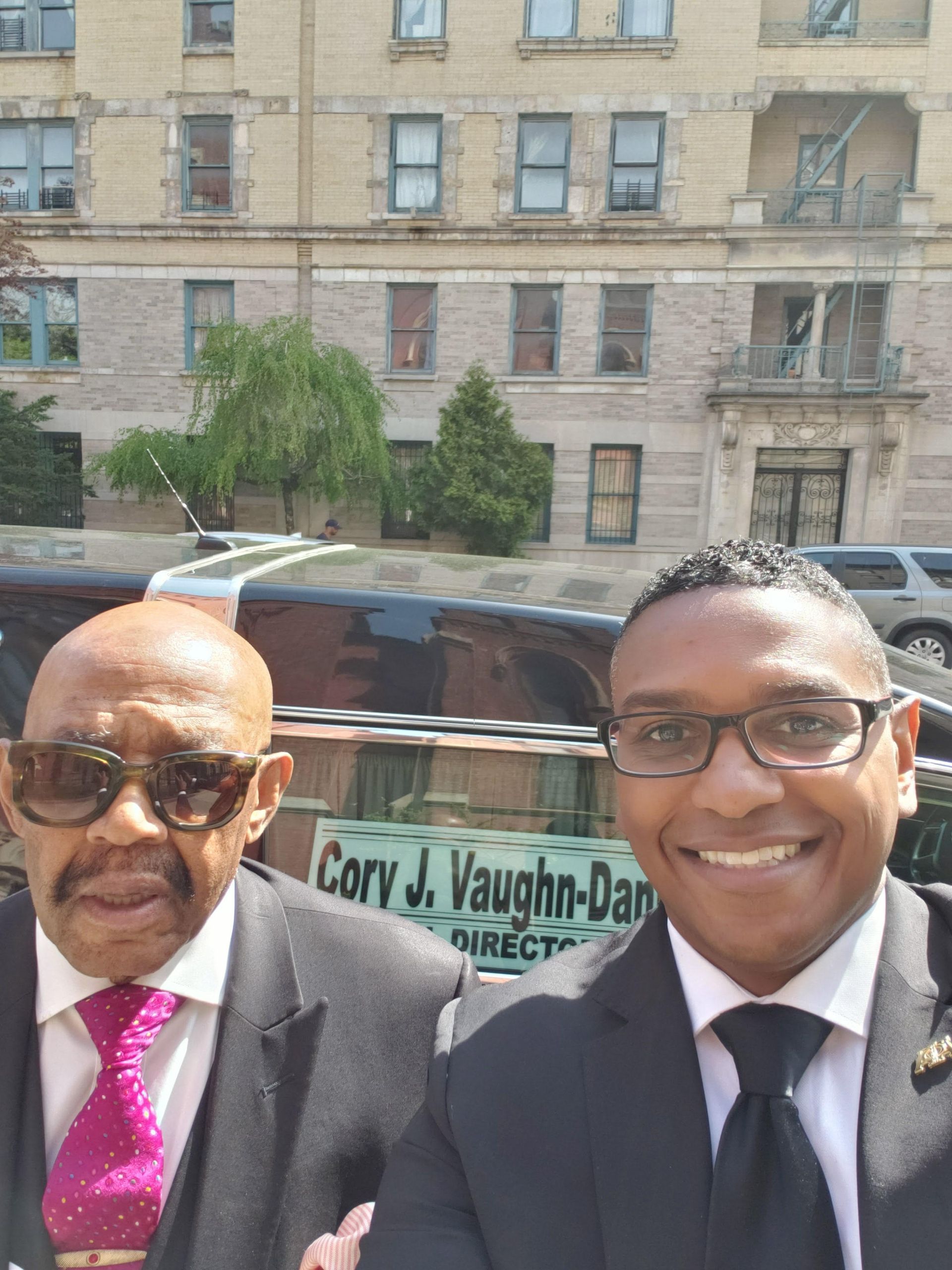 Two smiling men in suits posing outdoors in front of a building and a moving van.