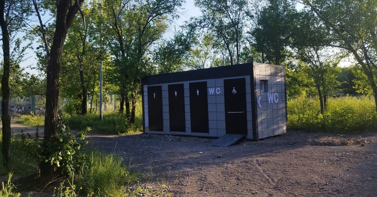 A modern portable restroom trailer rental sits in a wooded area. The unit features three individuals stalls and an ADA stall.