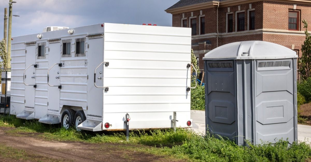 A gray portable toilet unit sits next to a white restroom trailer on set. The restroom trailer features multiple stalls.