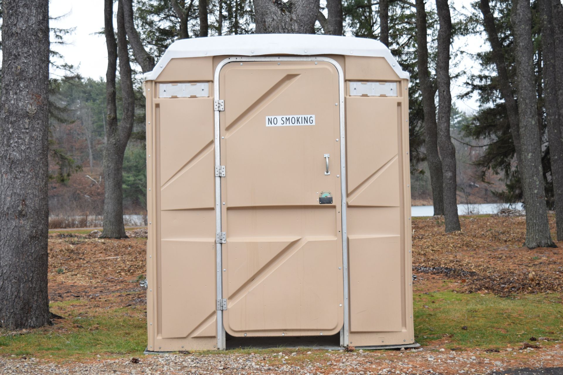 Beige porta potty in forest with “no smoke” sign, representing emergency response for porta potties. Beige porta potty in forest with “no smoke” sign, representing emergency response for porta potties.