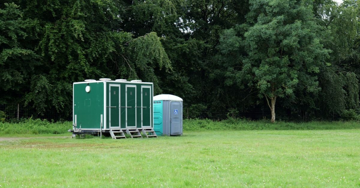 A restroom rental trailer sits in a field with three doors and steps up to the doors. A single unit stands next to it.