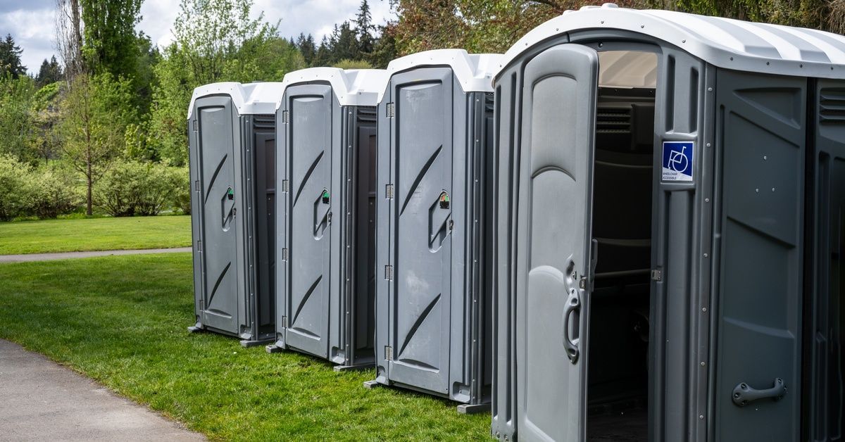 A row of portable toilet units sit on the grassy lawn next to a sidewalk. There are four ADA-complia