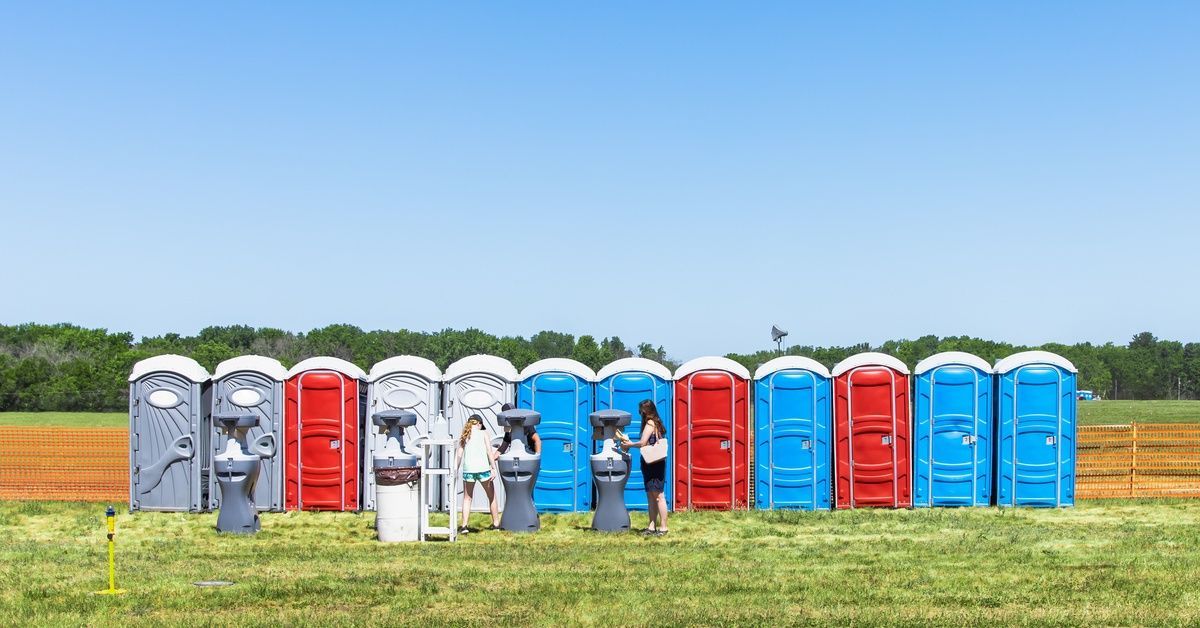 A row of blue, red, and gray portable toilet units in a field with handwashing stations in front and