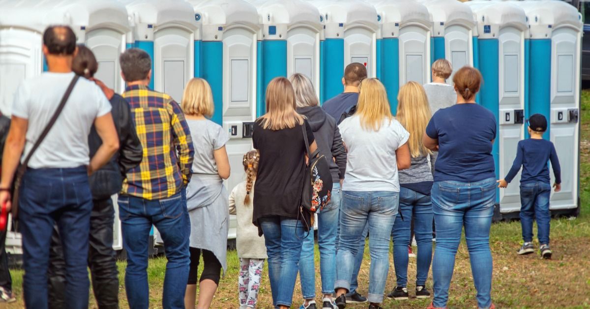 A long line of event attendees wait outside the portable restroom area. It's sunny outside and people are dressed casually.