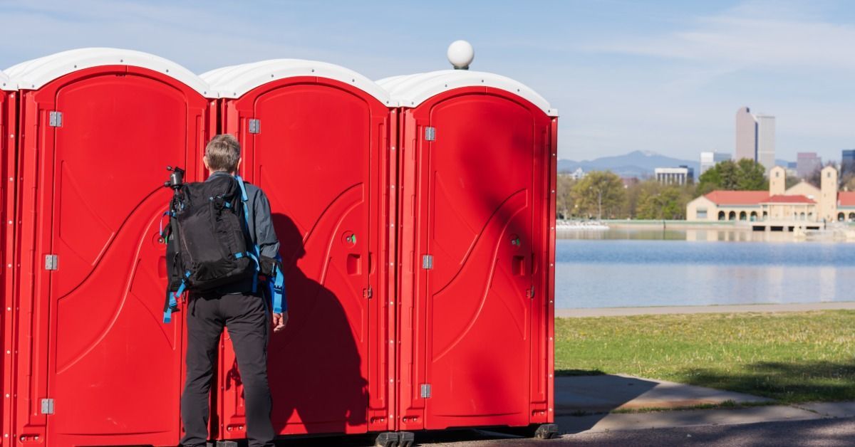 Three red portable restroom units sit together on a sidewalk near a river. There is a guest pulling the handle to use a unit.