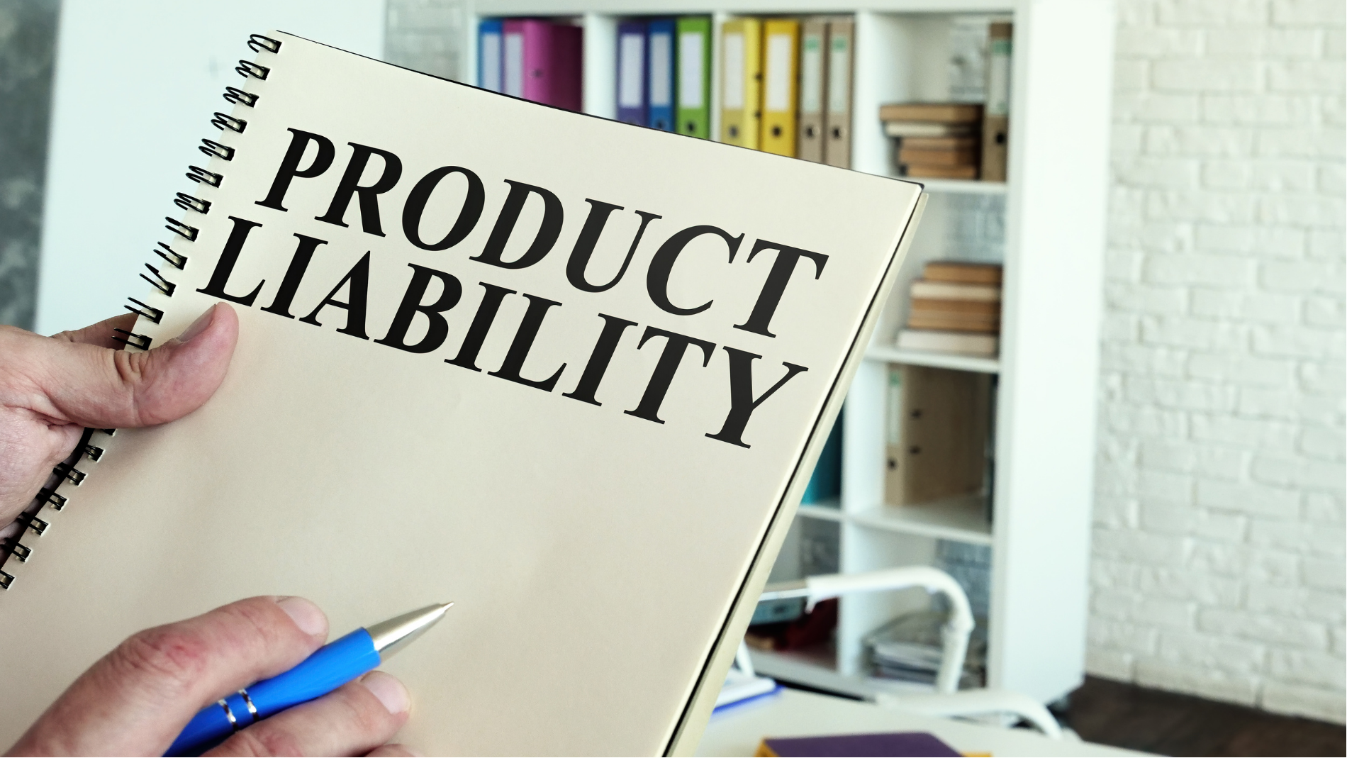 Hand holding a notebook labeled “PRODUCT LIABILITY” in an office setting