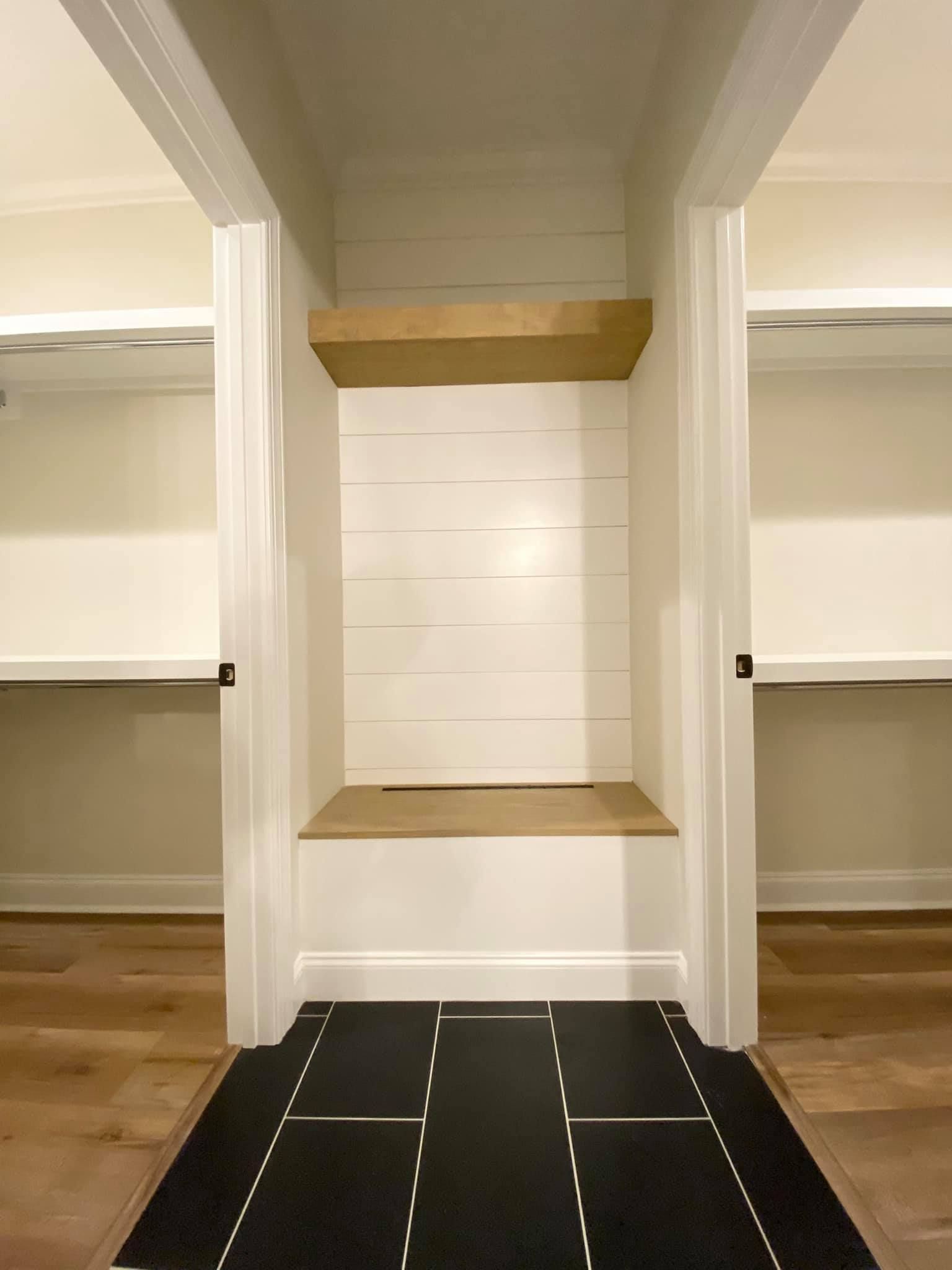 Closet hallway with built-in bench. White walls, wooden shelf and bench. Black and white patterned floor rug.