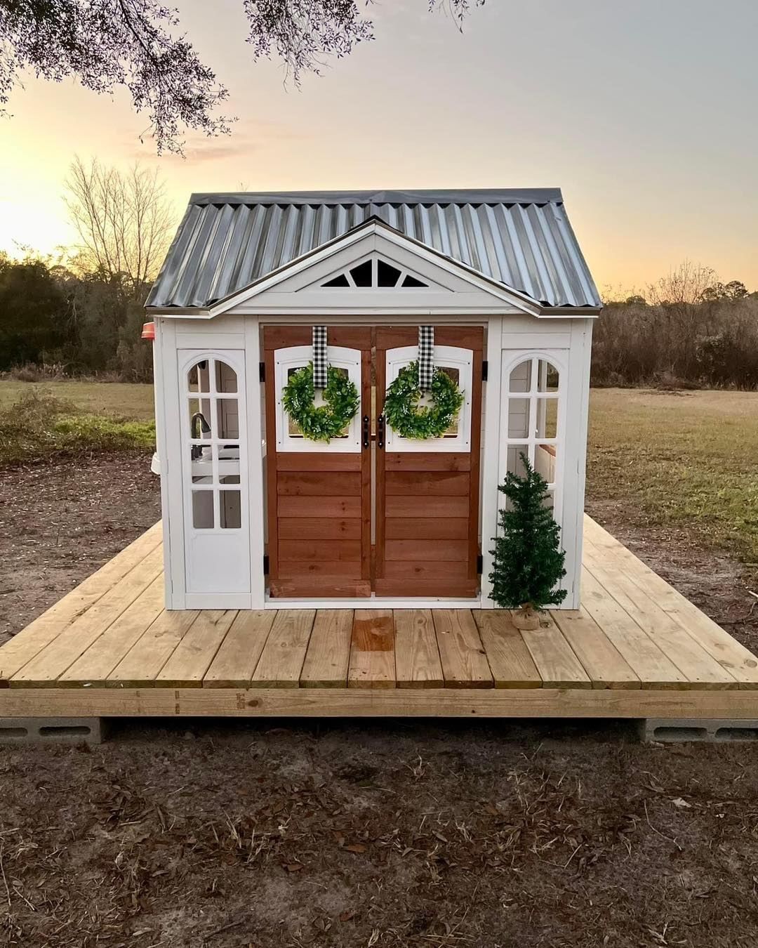 White playhouse with brown doors, green wreaths, on a wooden deck. Metal roof, small Christmas tree.