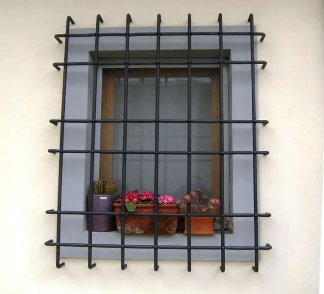 Finestra con inferriate di sicurezza in ferro nero e piccoli vasi di fiori sul davanzale, contro una parete beige.