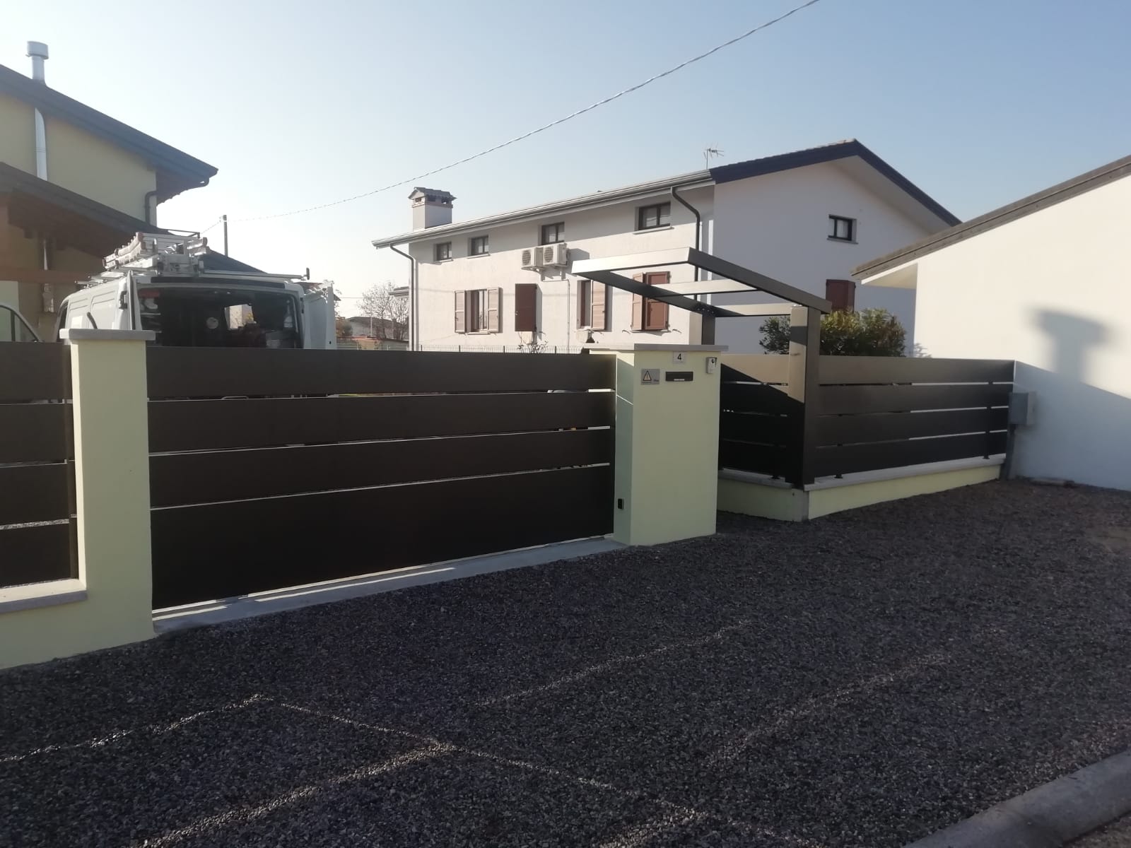 Esterno di una casa moderna con recinzione scura, cancello e vialetto di ghiaia sotto un cielo limpido