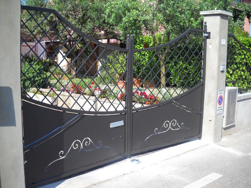 Cancello in metallo nero con motivo a griglia all'ingresso di un'abitazione, con giardino e pilastri in pietra alle sue spalle.