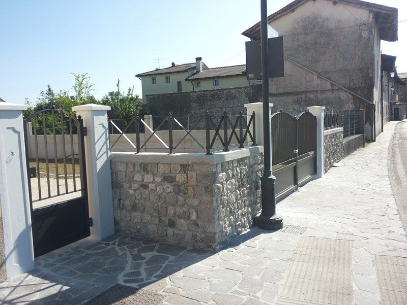 Muro di pietra e cancello nero accanto a un marciapiede soleggiato e a una casa rustica in una strada tranquilla.