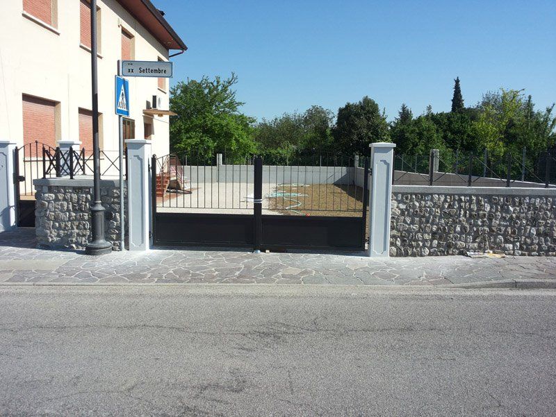 Cancello residenziale con pilastri in pietra e recinzione in metallo nero accanto a una casa in una strada soleggiata