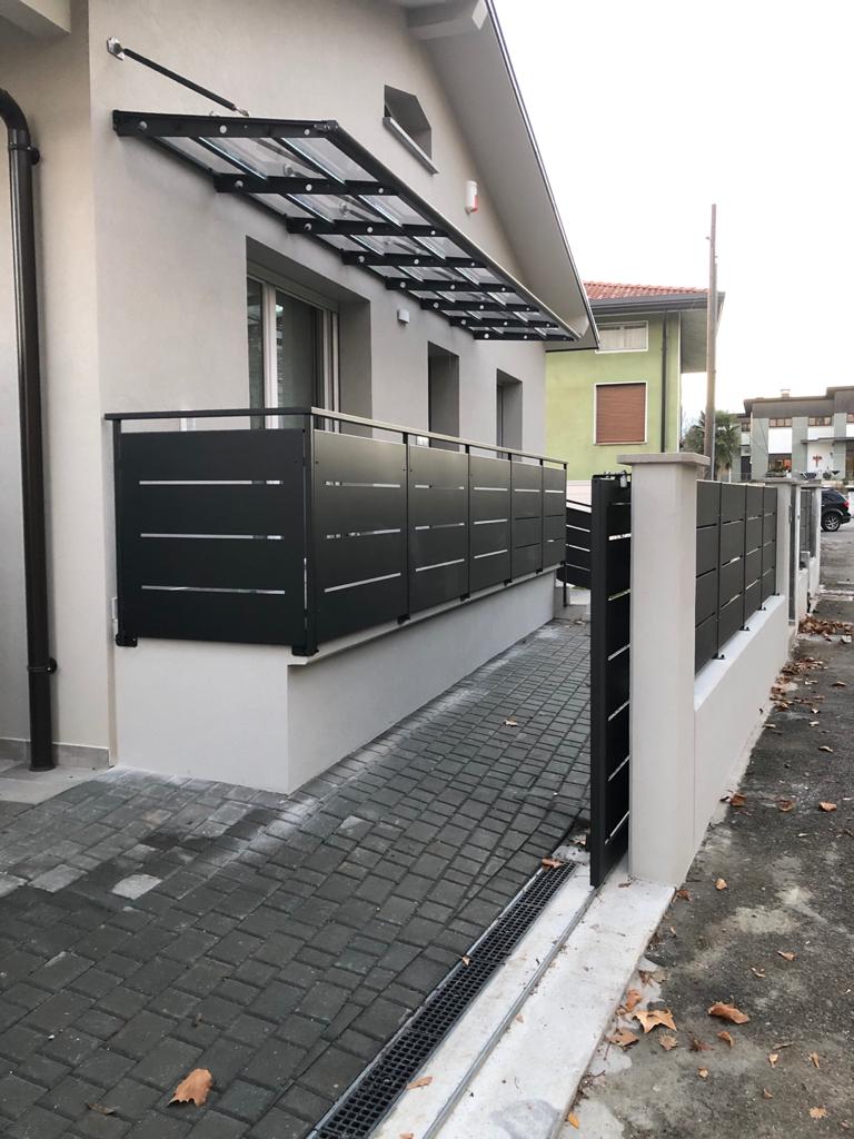 Ingresso moderno della casa con cancello nero, vialetto pavimentato e tettoia metallica sul sentiero laterale.