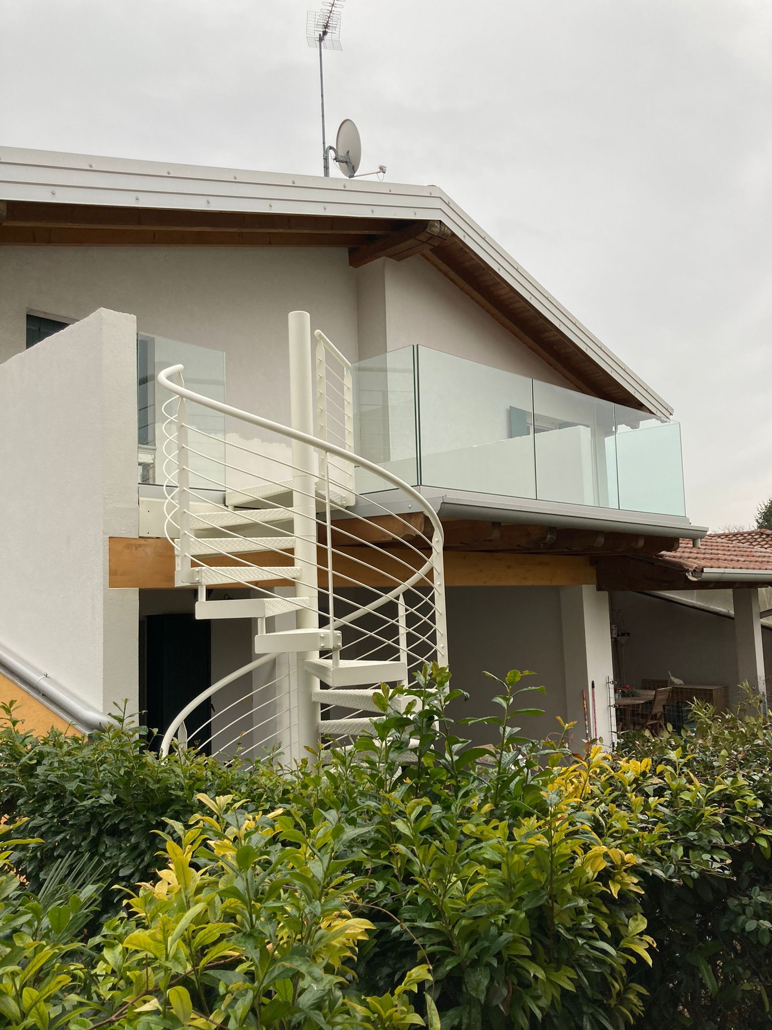 Esterno di una casa moderna con scala bianca curva, balcone in vetro e arbusti verdi sul davanti.