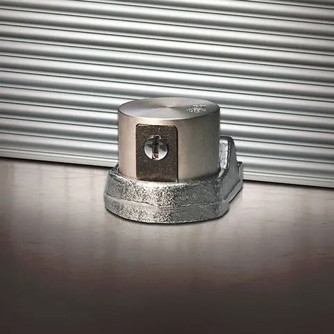 Tappo esagonale metallico su base argentata, appoggiato su un piano riflettente davanti a una porta in lamiera ondulata.