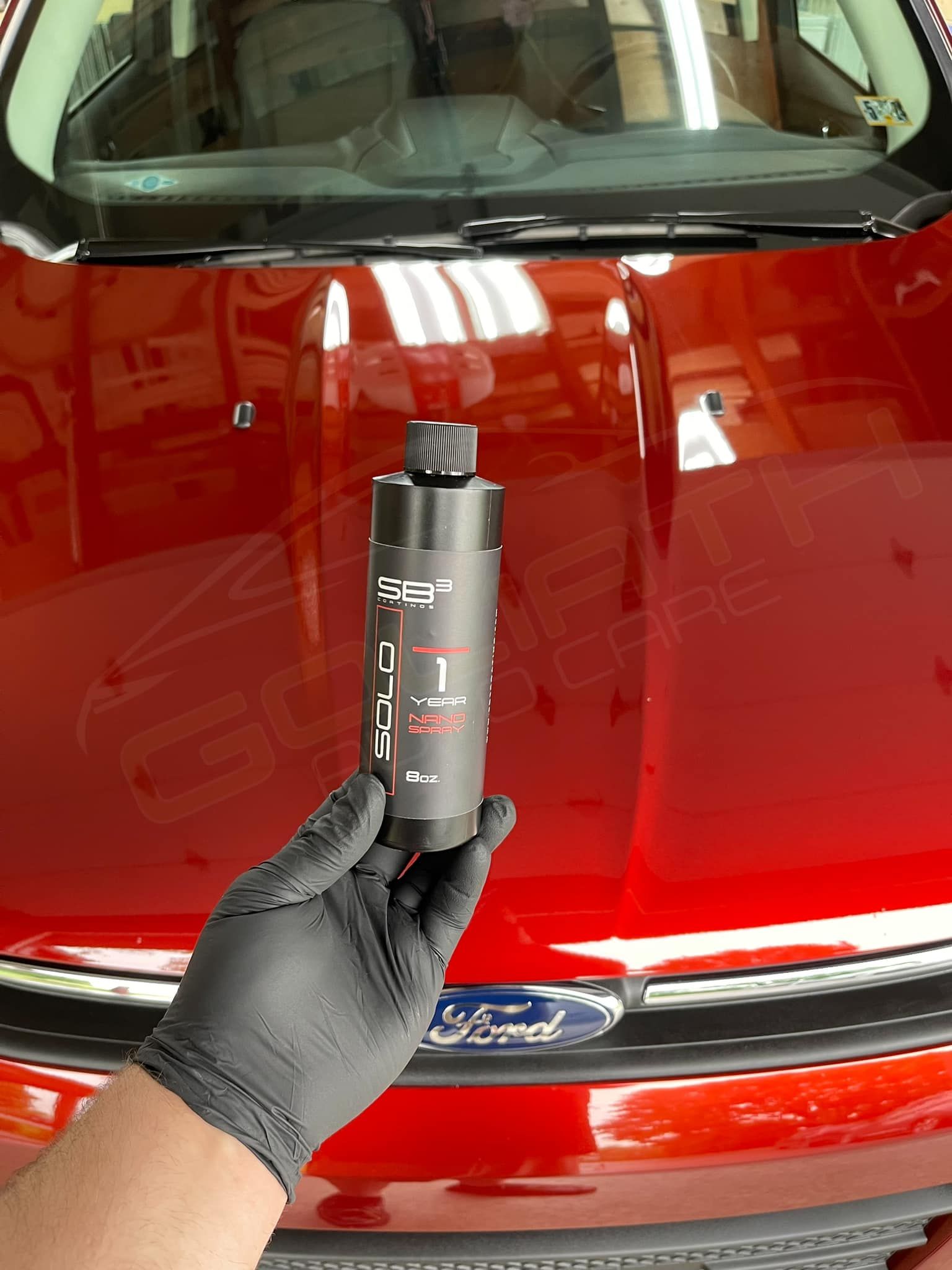 a person wearing black gloves is holding a bottle in front of a red car .