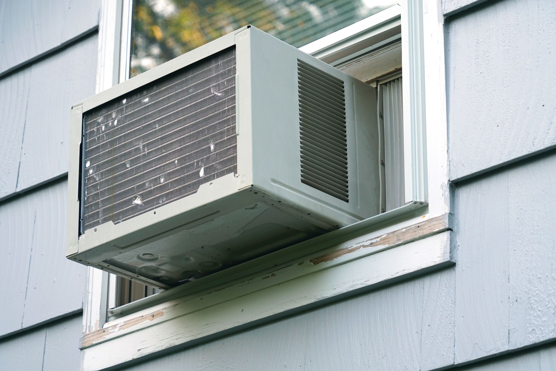 Window air conditioner mounted in a house window, with a gray exterior wall.