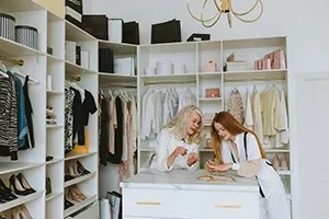 Two people browsing clothes in a bright white walk-in closet with shelves, hanging garments, and shoes
