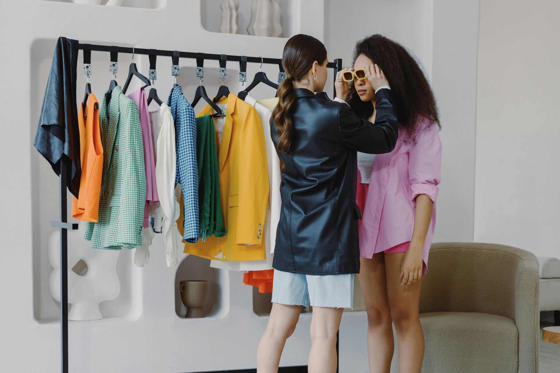 Two women browsing clothes on a rack in a bright room, one holding up sunglasses