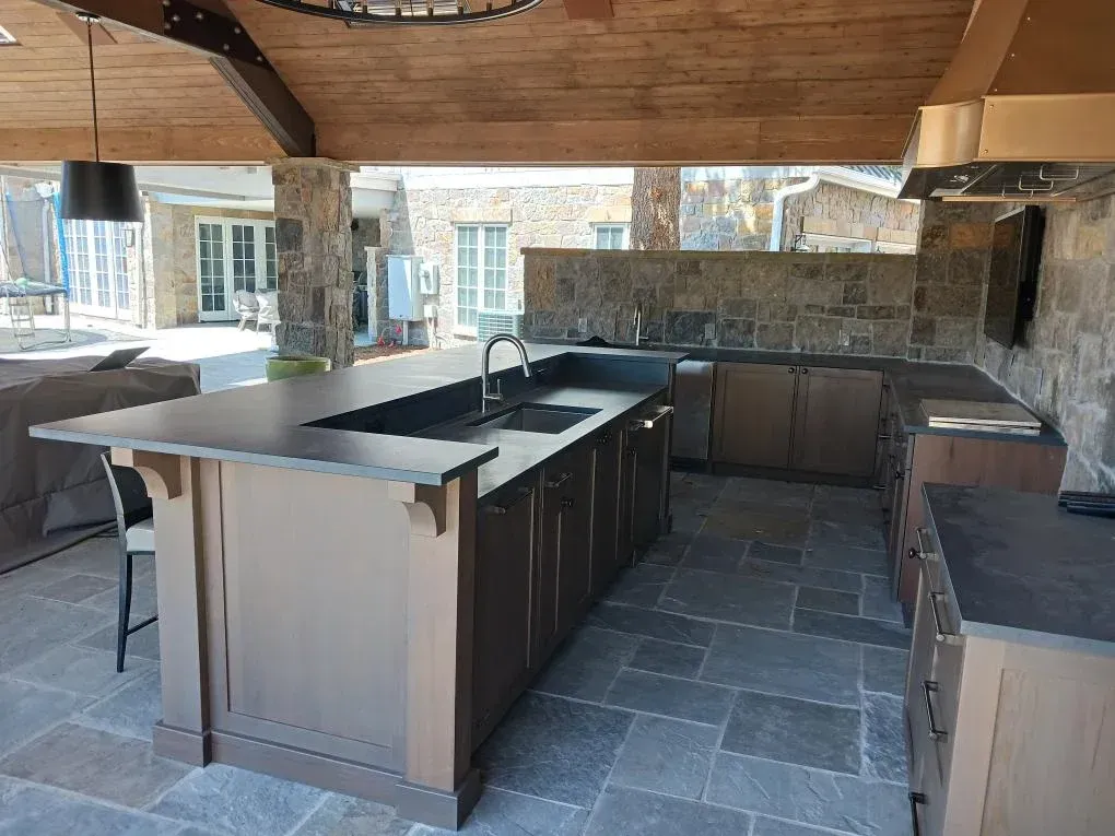 Outdoor kitchen with large granite island, built-in grill, and bar seating under a wooden pergola.