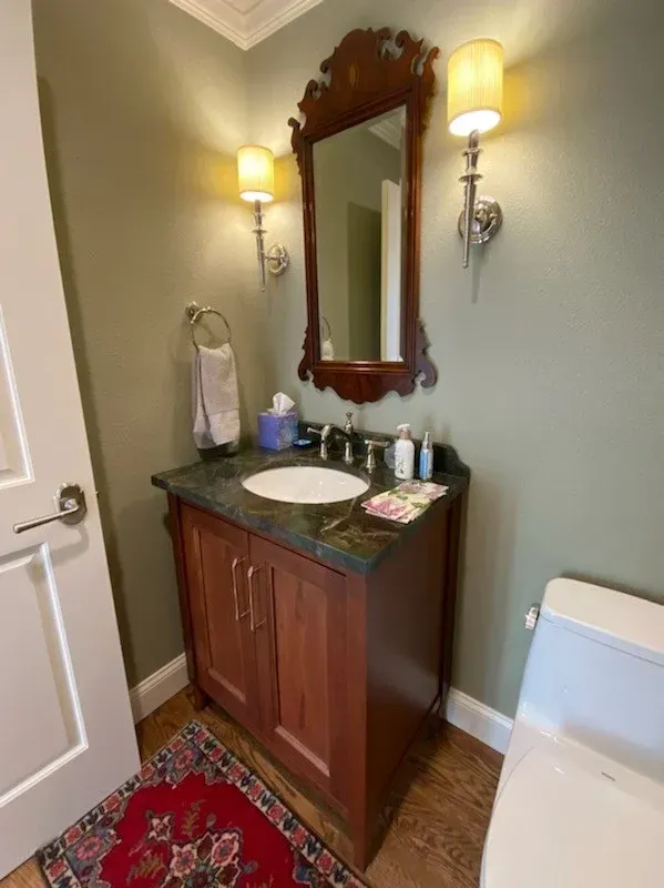 Small bathroom vanity with mirror, wall sconces, red rug, and toilet in a narrow corner.