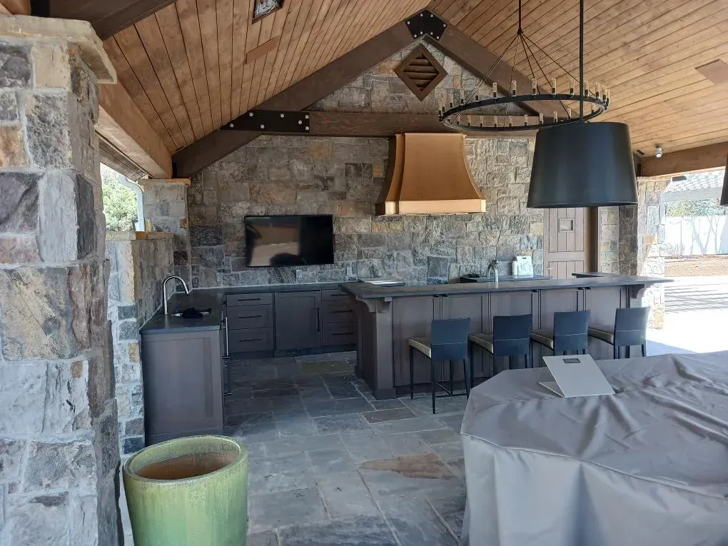 Rustic outdoor kitchen with stone bar, wood ceiling, grill, stools, and covered table