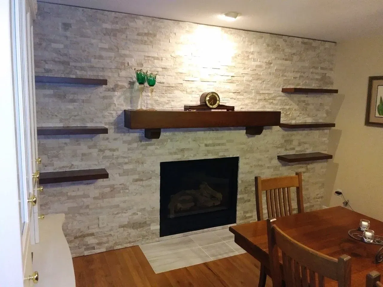 Living room fireplace with stone surround, wood mantel, wall shelves, and a dining table in the foreground