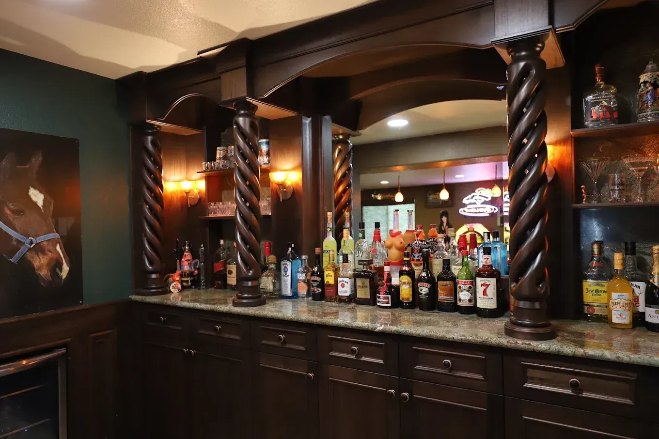 Dim bar with dark wood cabinetry, twisted columns, and a counter lined with liquor bottles.
