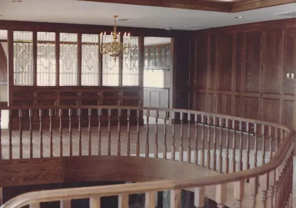 Empty wooden courtroom gallery with curved railing, chandeliers, and windows along the back wall
