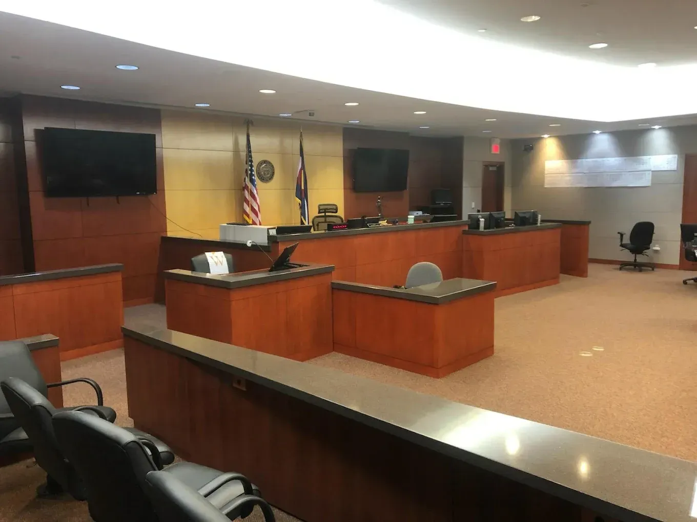 Empty courtroom with wooden benches, judge’s bench, flags, and flat-screen monitors under bright ceiling lights