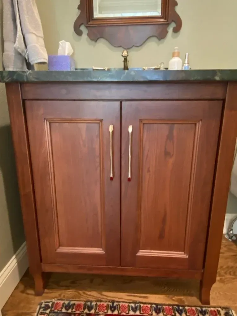 Wooden bathroom vanity with dark countertop, double cabinet doors, and sink accessories on top