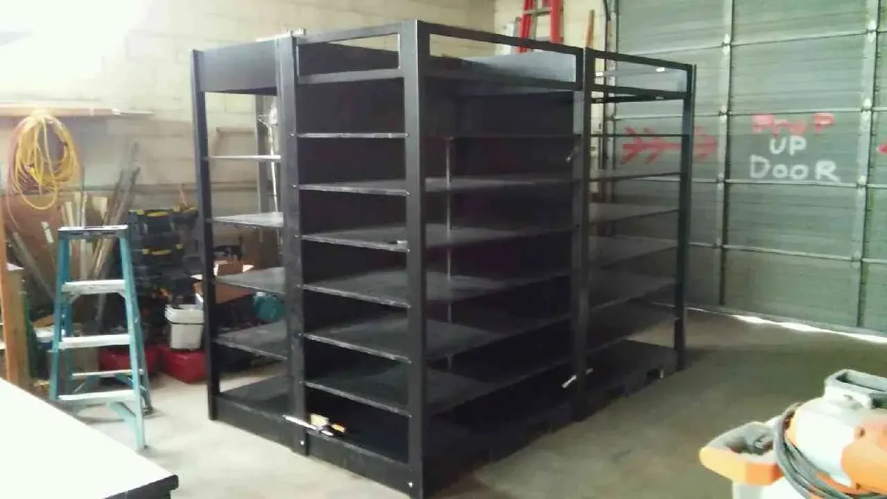Black metal shelving racks in a workshop or warehouse, with empty shelves and a red wall in the background.