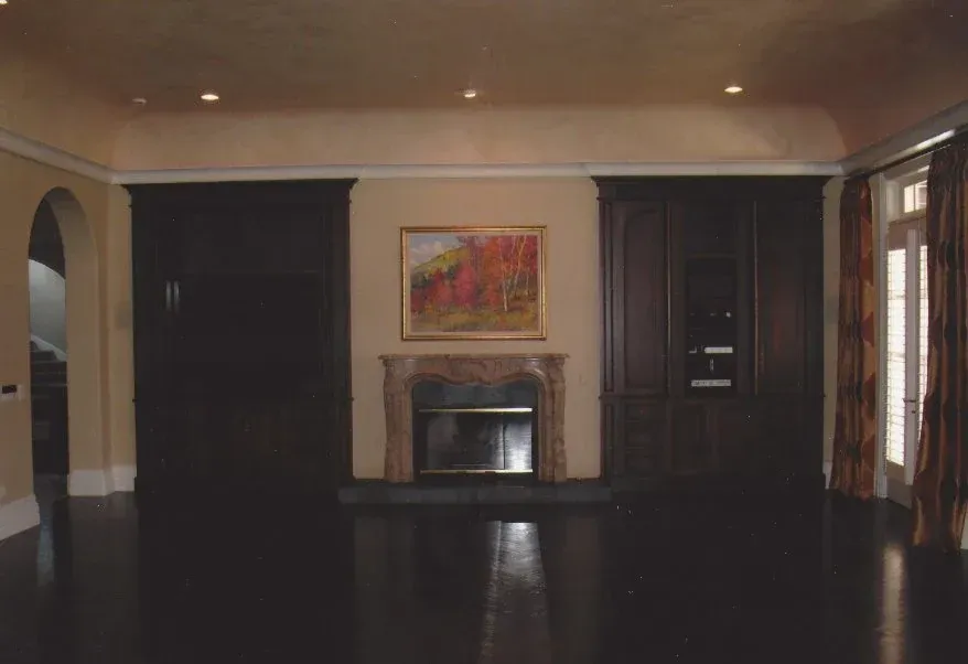 Empty living room with a fireplace, dark wood floors, beige walls, and curtains by tall windows