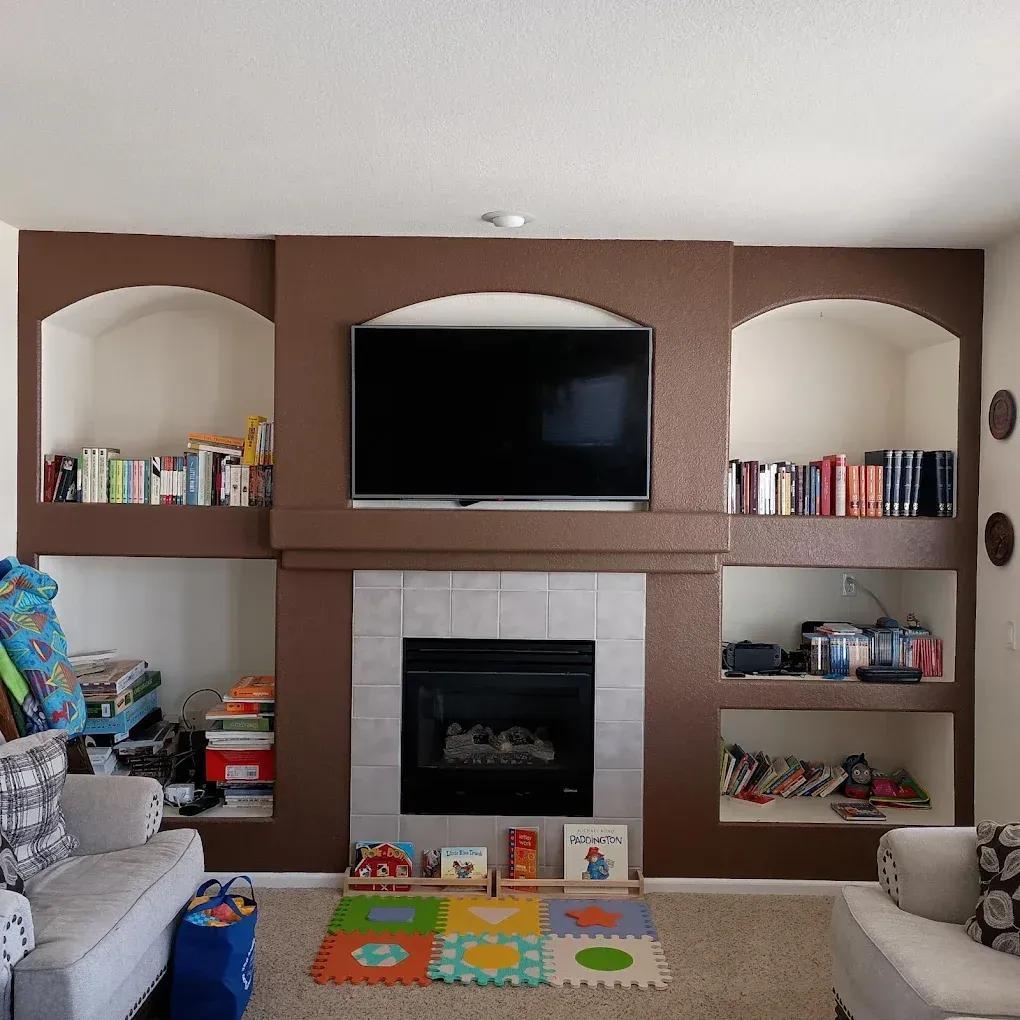 Living room with brown built-in shelves, a TV above a fireplace, and books and toys on display.